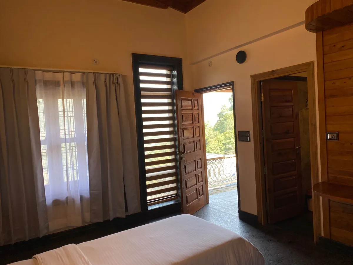 Ashram guest room with wooden shutters, soft curtains and a view of the hillside through the open door