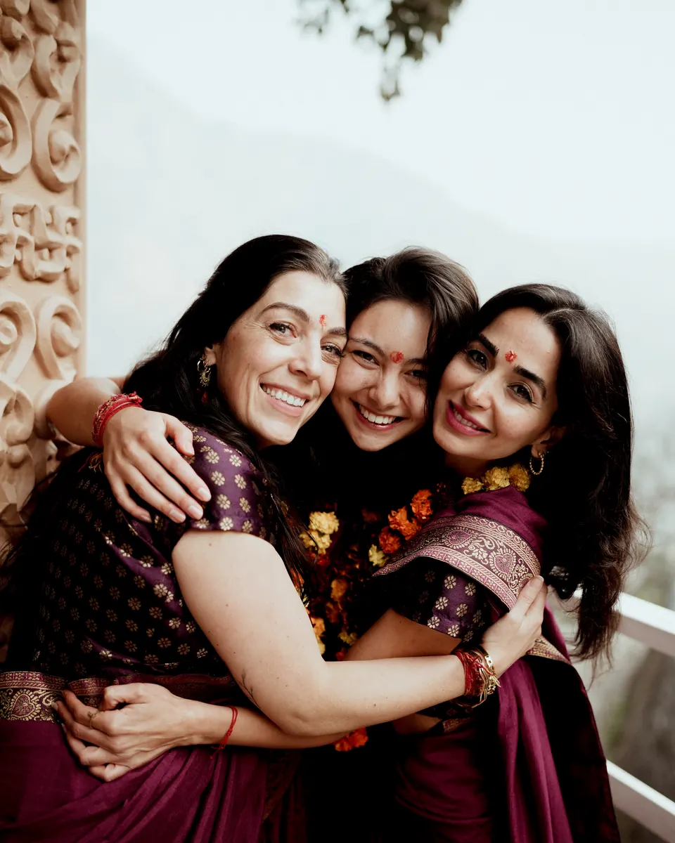 Three graduates in saris and marigold garlands hug and laugh on graduation day at the ashram