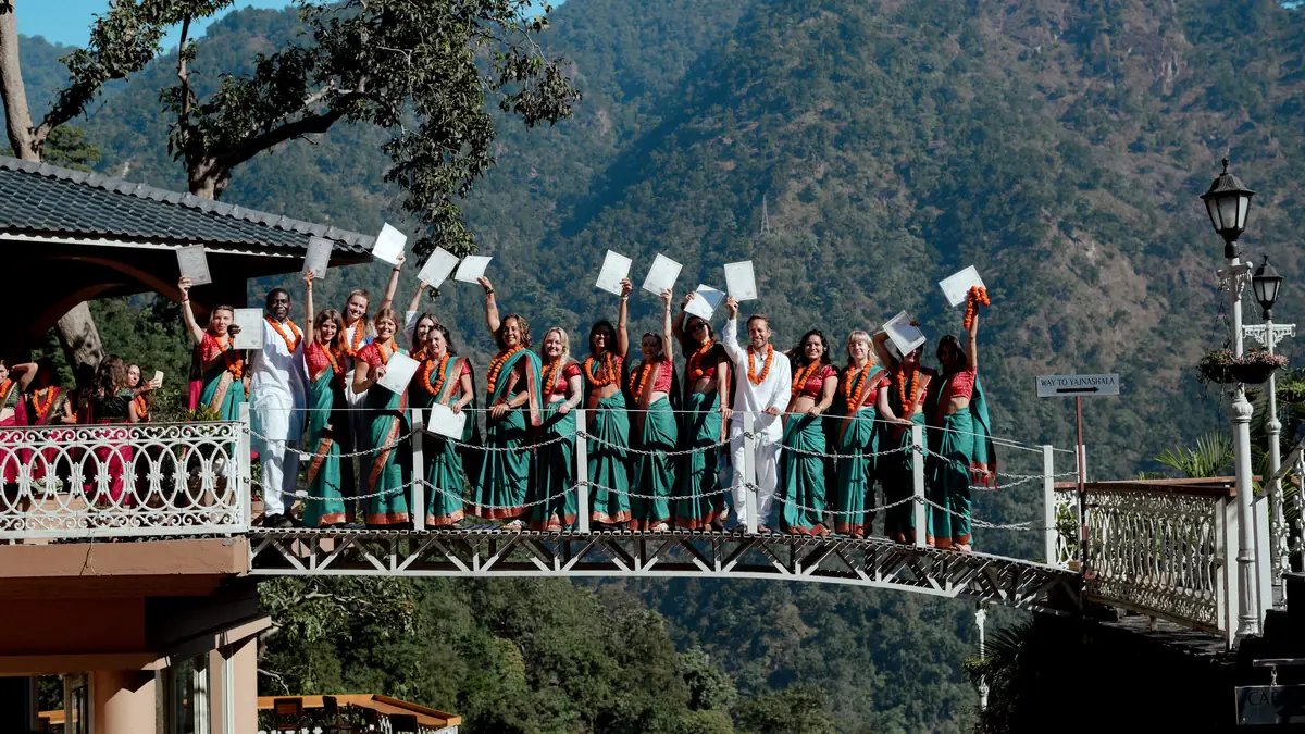 Students from many countries wave their certificates on the ashram bridge beneath a blue Himalayan sky
