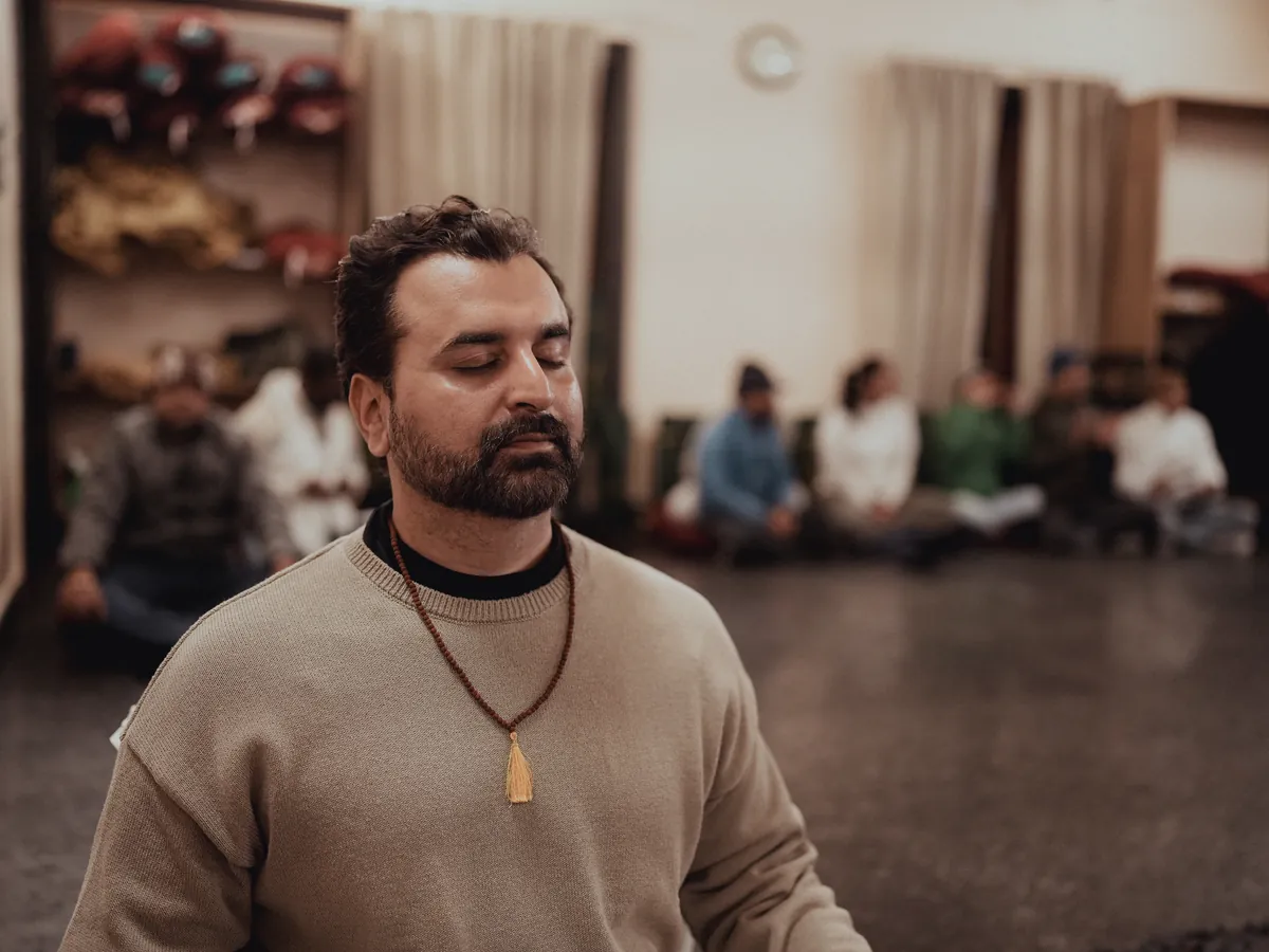 A teacher wearing rudraksha beads sits in meditation in front of students in the yoga hall