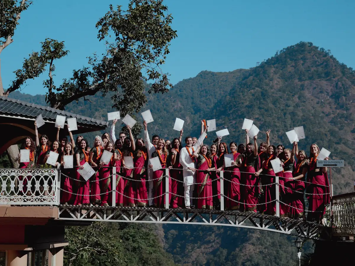 A teacher training batch in red saris and marigold garlands holds up their certificates on the ashram bridge, with forested mountains behind