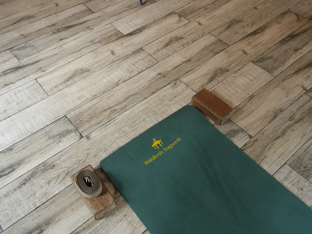 A green Rishikesh Yogpeeth yoga mat with wooden blocks and a cotton strap on the floor of the yoga hall