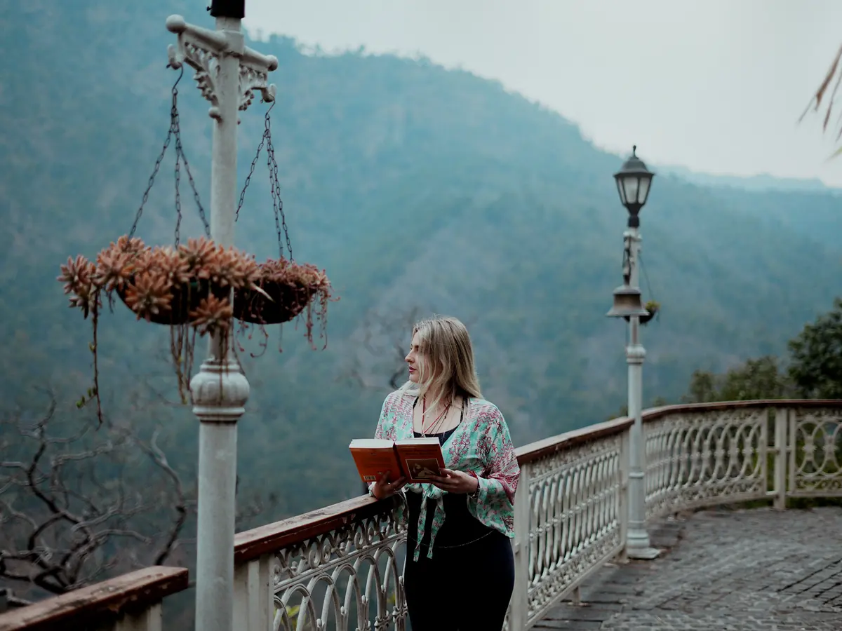 A student stands at the railing of an ashram terrace with a book in her hands, looking out over the forested hills near Rishikesh